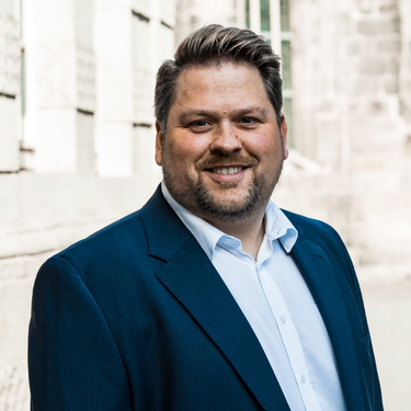 Mann mit dunklem Sakko und offenem Hemd steht lächelnd vor einem alten Stein-Gebäude.