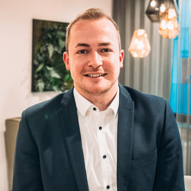 Junger Mann im Sakko steht in gemütlich beleuchtetem Raum und blickt lächelnd in die Kamera.