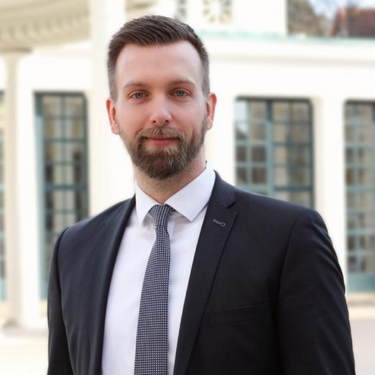 Mann mit Bart im Anzug steht vor einem Gebäude mit weißen Säulen und Fensterfront.