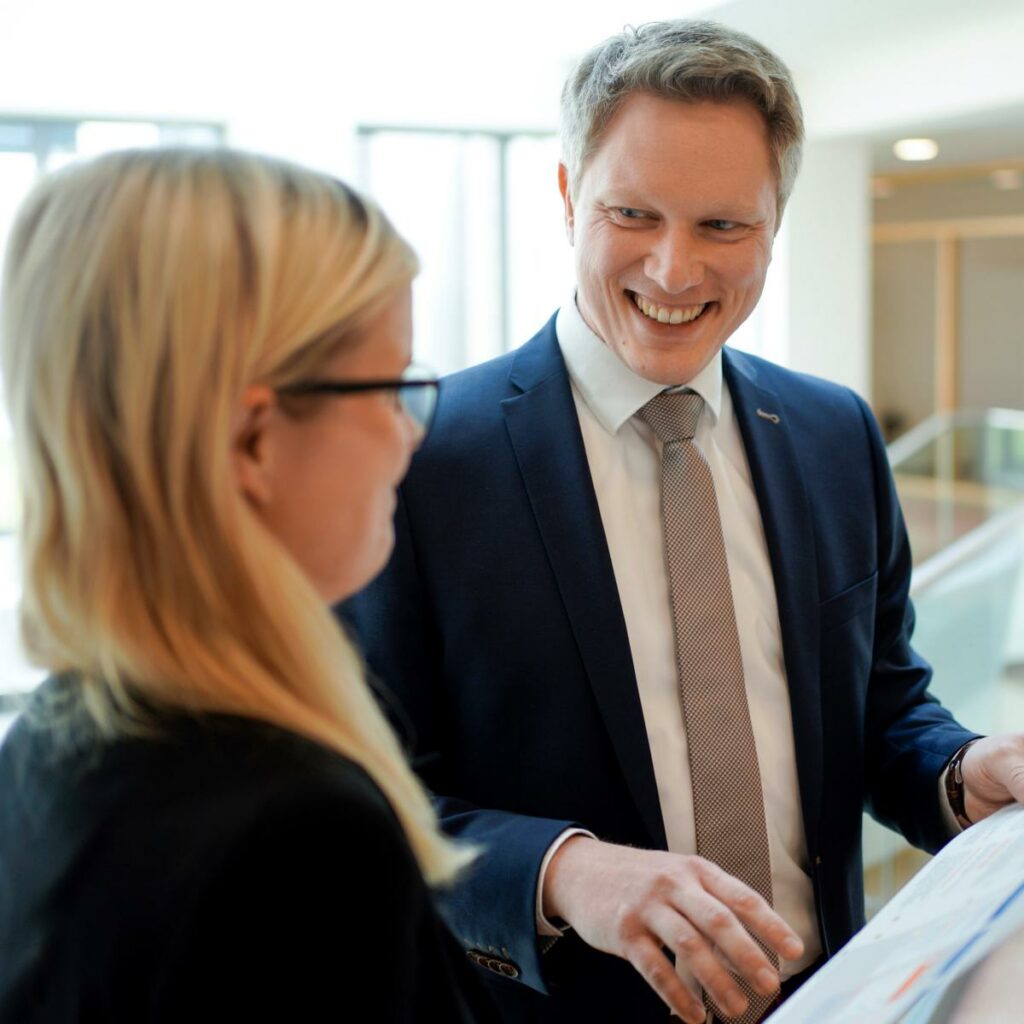 Mann im Anzug zeigt einer Kollegin etwas auf einem Papier und lächelt dabei. Beide stehen in einem modernen Innenraum.