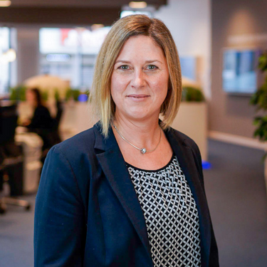 Frau mit blondem Bob und Blazer steht in einer modernen Büroumgebung und blickt freundlich in die Kamera.