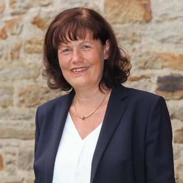 Frau mit schulterlangen dunklen Haaren im Blazer lächelt vor einer Natursteinwand.