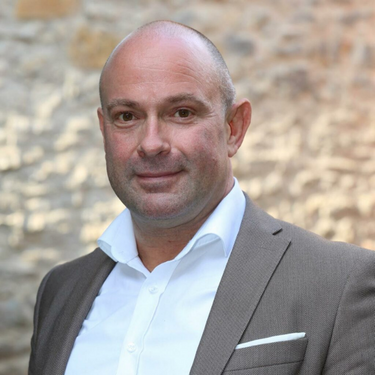 Mann mit Glatze im hellbraunen Anzug und weißem Hemd steht vor einer Natursteinwand und blickt freundlich in die Kamera.