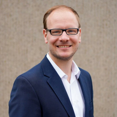 Mann mit Brille und Anzug lächelt freundlich vor einer schlichten Wand.