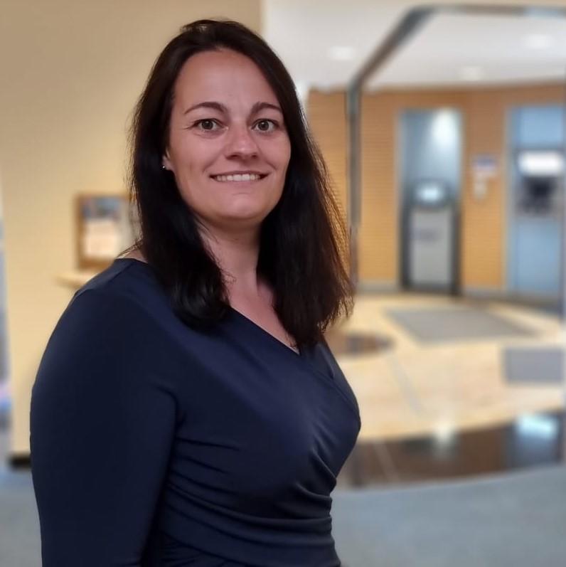 Frau mit dunklen Haaren steht lächelnd in einer Bankfiliale mit Automaten im Hintergrund.