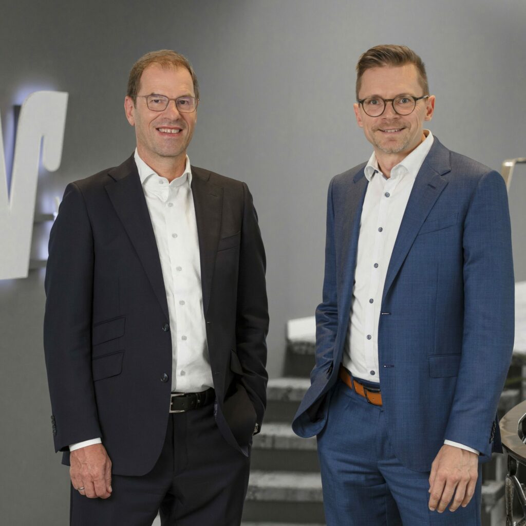 Zwei Männer in Anzügen stehen nebeneinander vor grauer Wand mit Logo, beide lächeln in die Kamera.