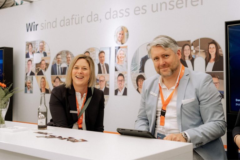 Eine Frau und ein Mann sitzen an einem Messestand mit Getränkeflasche und Tablet. Im Hintergrund sind Fotos von Teammitgliedern zu sehen.