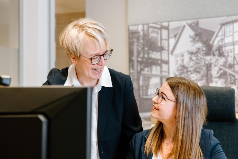 Zwei Frauen in Business-Kleidung unterhalten sich freundlich an einem Arbeitsplatz mit Computer. Eine Frau sitzt, die andere steht daneben.