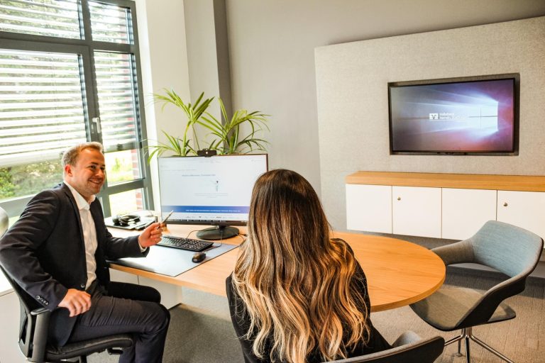 Ein Mann im Anzug berät eine Frau mit langen Haaren an einem runden Schreibtisch in einem hellen Büro.