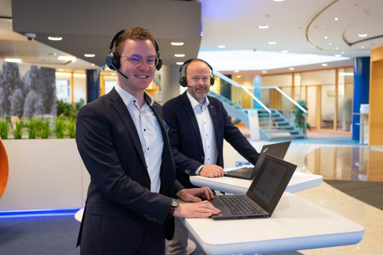 Zwei Mitarbeiter mit Headsets stehen mit Laptops an einem Stehtisch und lächeln.