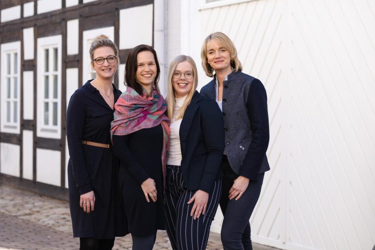 Vier Frauen stehen nebeneinander und lächeln in die Kamera. Sie tragen Business-Kleidung und stehen vor einem traditionellen Fachwerkhaus.