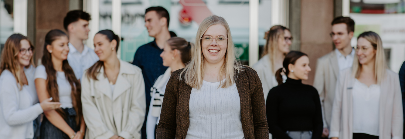Junge Frau mit Brille steht lächelnd im Vordergrund, im Hintergrund unterhalten sich mehrere junge Menschen in lockerer Kleidung.