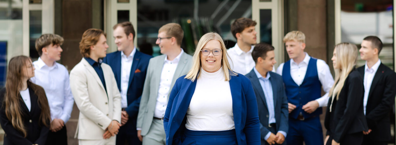 Junge Frau mit Brille steht lächelnd im Vordergrund, im Hintergrund unterhalten sich mehrere junge Menschen.
